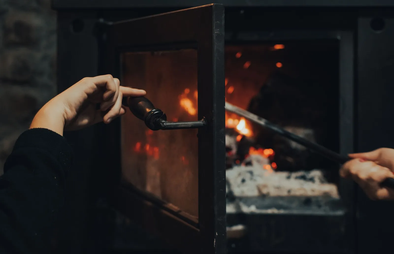 Main ouvrant la porte vitrée d'un poêle à bois devant un feu de bûches et un lit de braises